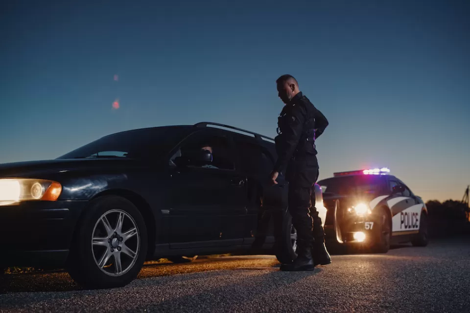Police office standing next to sedan he just pulled over at night with police car in background.